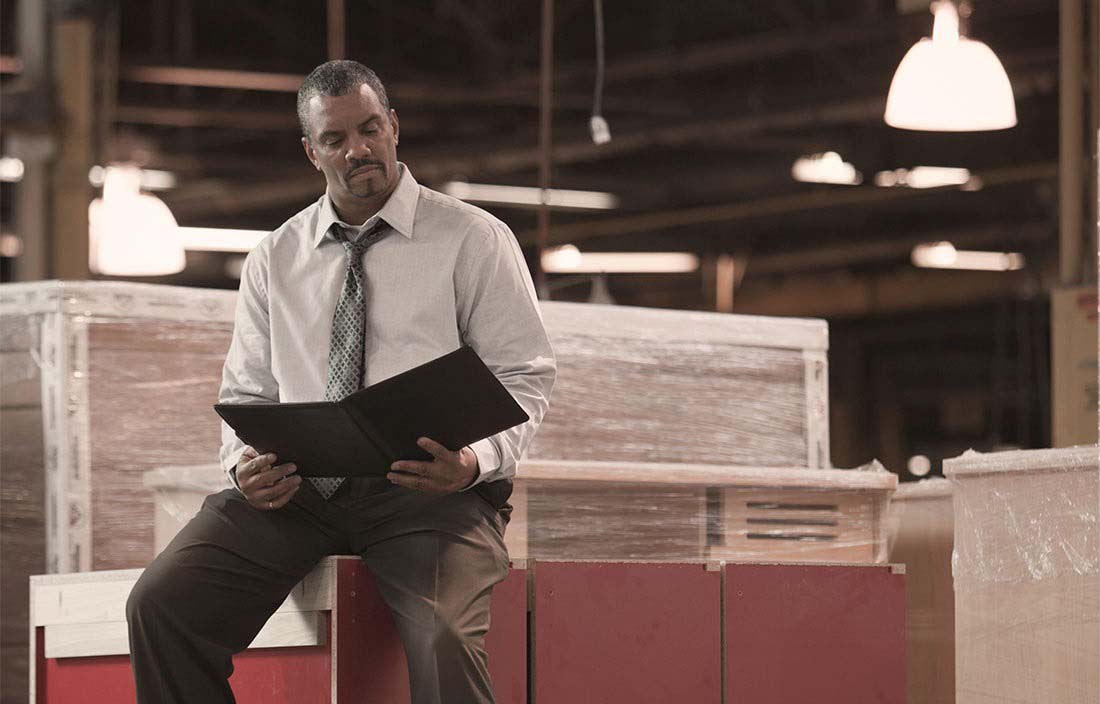 Image of a man sitting on a box in a warehouse, reading from a book.