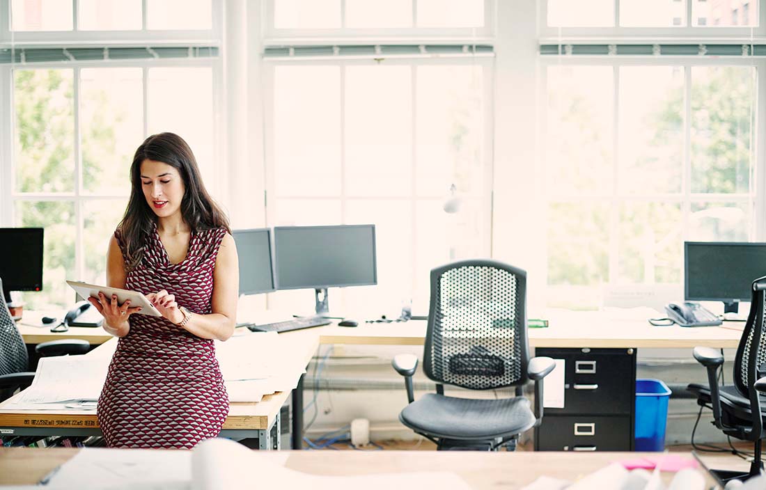 A woman in a modern office setting reading news on a tablet handheld device. 