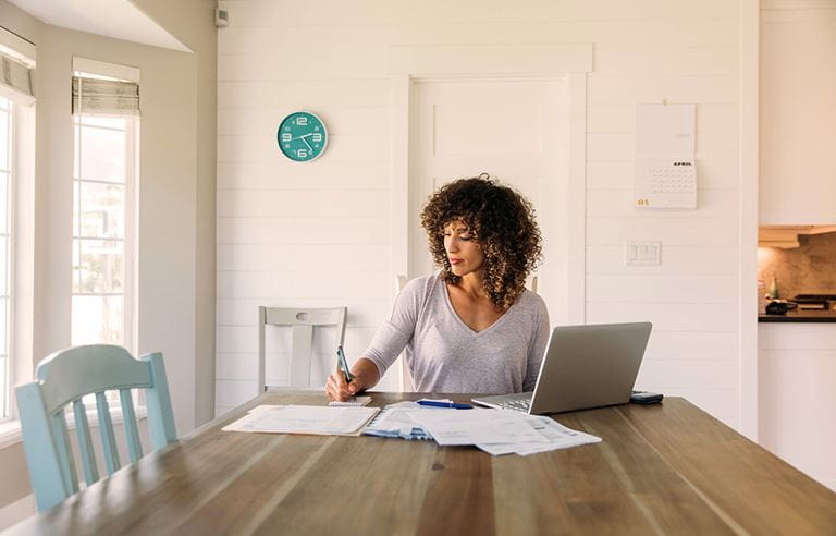 Woman sitting at a kitchen table writing on notepad with her laptop computer as well.