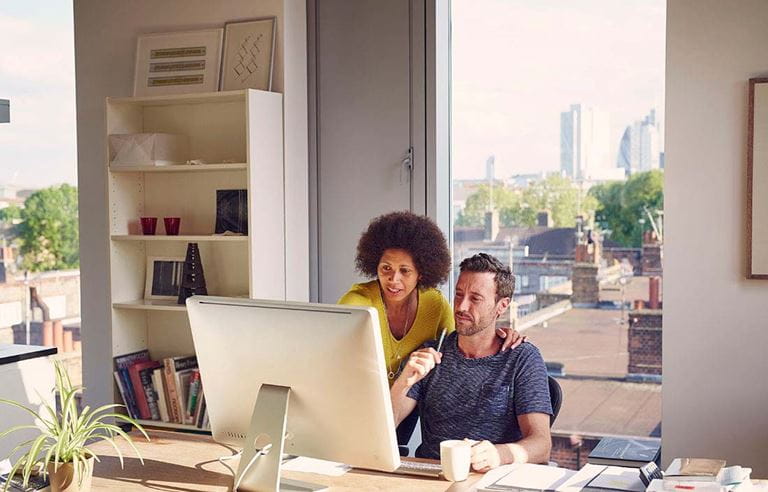 A man and woman sitting at a desktop computer in a personal office looking at the monitor screen.