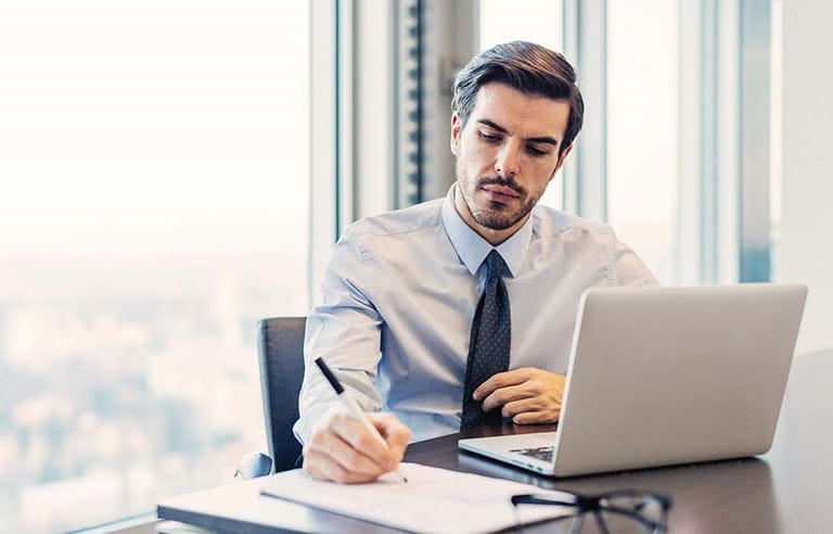 Business professional working at a desk writing on a document with his laptop computer out.