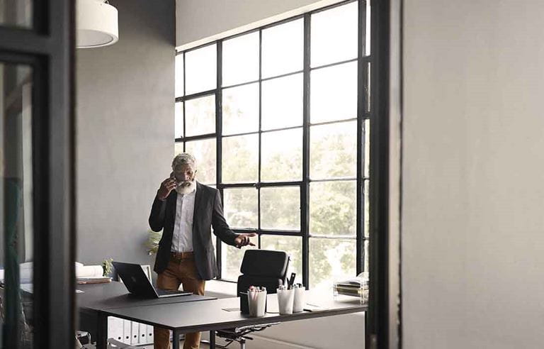 Elderly businessman taking a phone call at home standing by his desk.