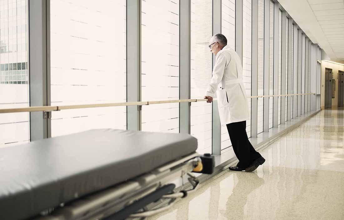 Doctor in a hallway leaning against a railing looking out the window.