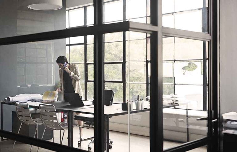 Businessperson standing in an office reviewing documents on a desk while taking a phone call.