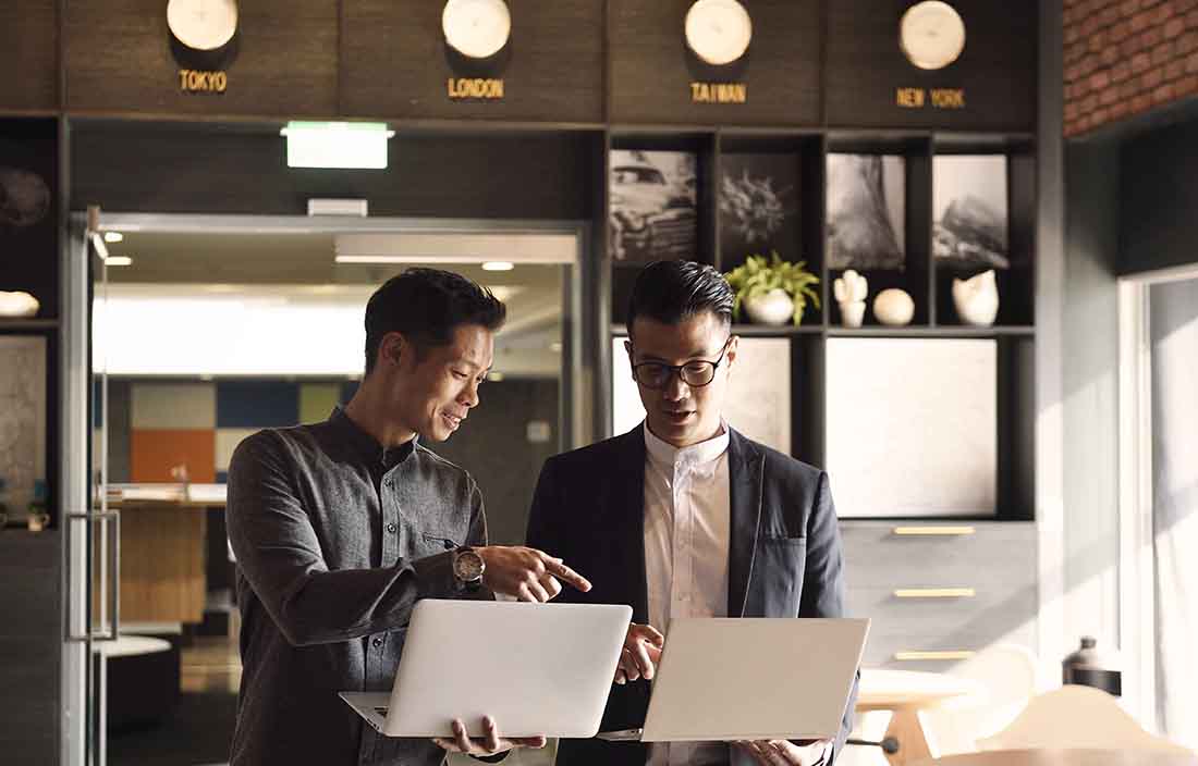 Two business professionals holding laptops while discussing amongst each other.