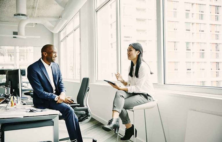 Two business professionals sitting by a desk having a conversation while smiling.