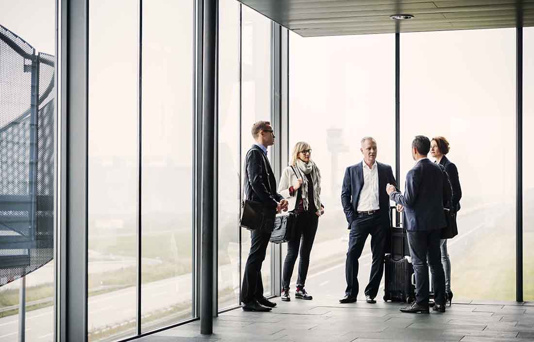 Five business professionals standing in the corner of a meeting room talking.