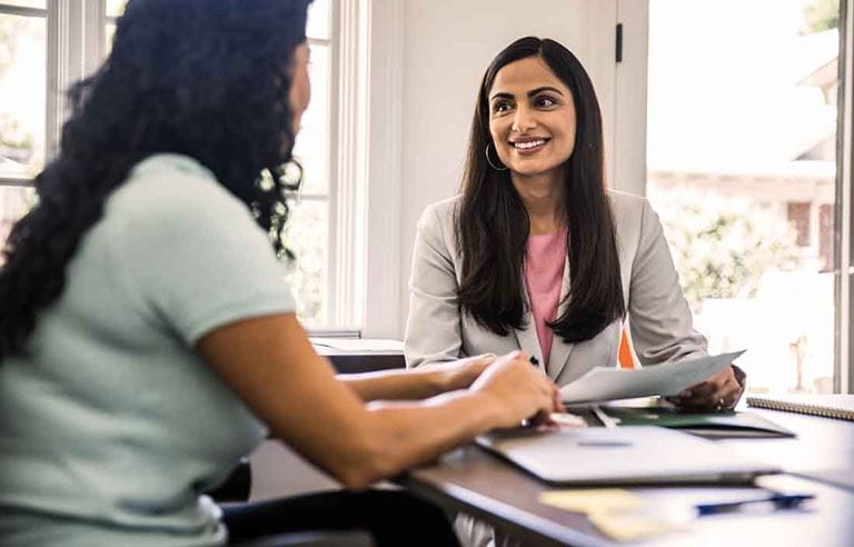 Two business professionals smiling and talking with one another while sitting at a desk.