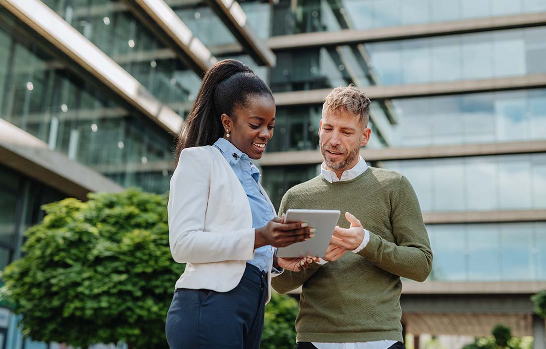 Two business professionals in casual clothing using a handheld tablet device together while standing.