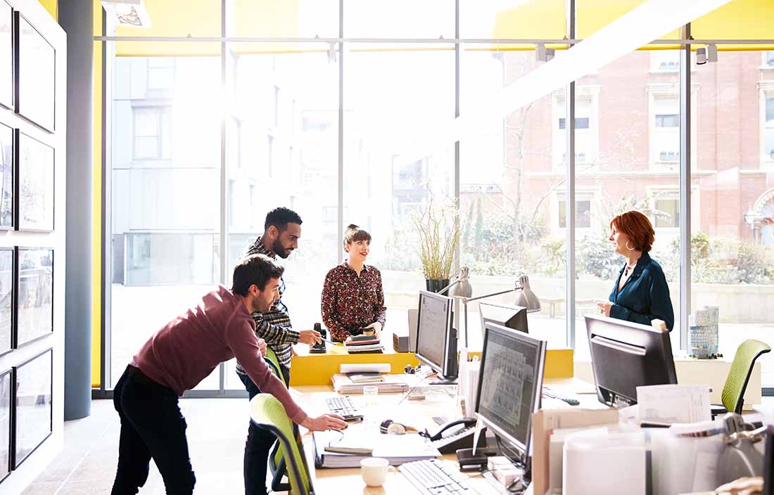 Coworkers at a nonprofit gathered around their desks and chatting in a sunny room.