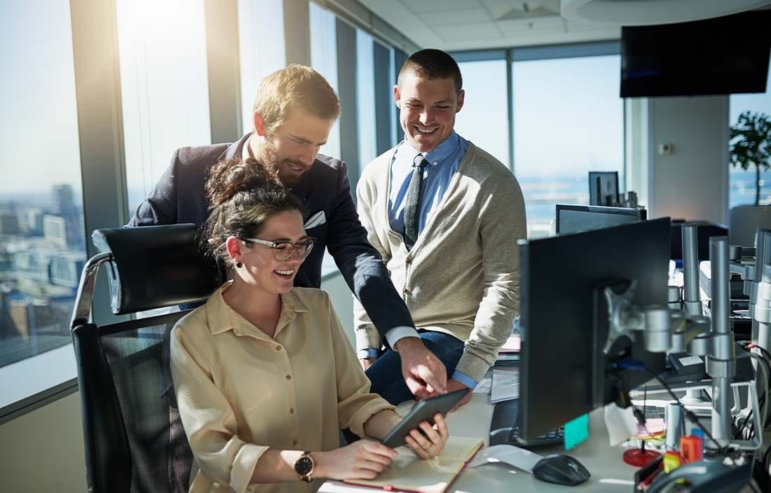 Business professionals huddled around a computer smiling while reviewing information on a tablet device