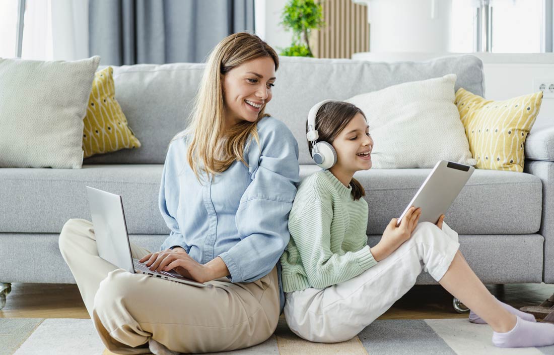 Parent and child sitting back-to-back on their living room floor on their laptop and tablets.