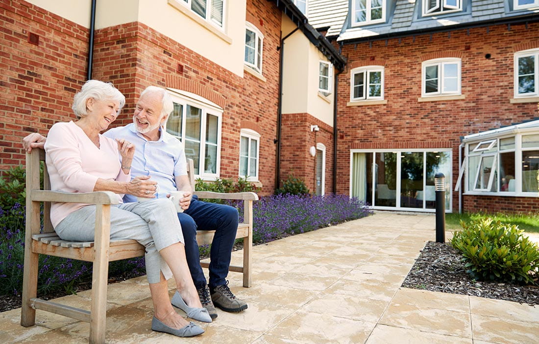 Two seniors at a senior living community on a bench in the foreground with the building in the background