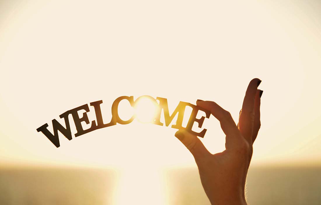 Hand holding up a wooden sculpture that says "welcome" against a sunrise or sunset light