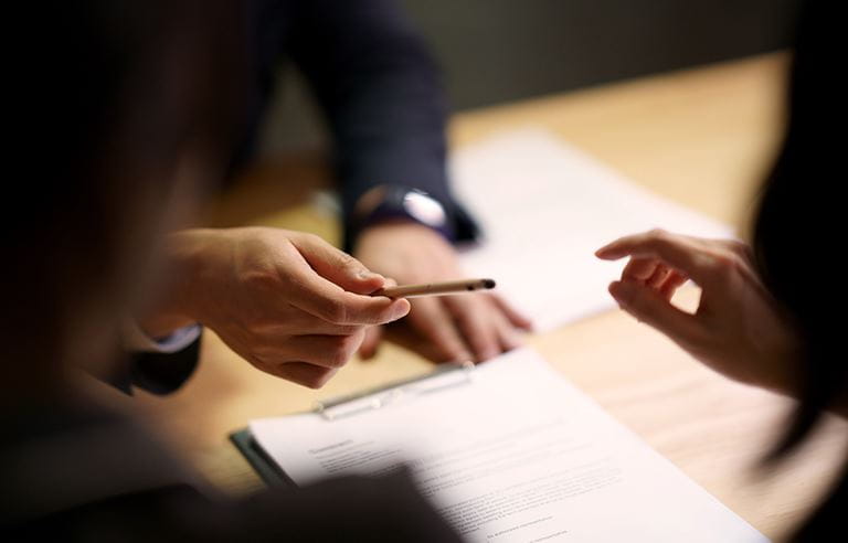  Looking through the shadowy figures of two businesspeople, a hand of another businessnesperson is giving a pen to one of them, who is reaching for it in order to sign contract agreement on the table between them