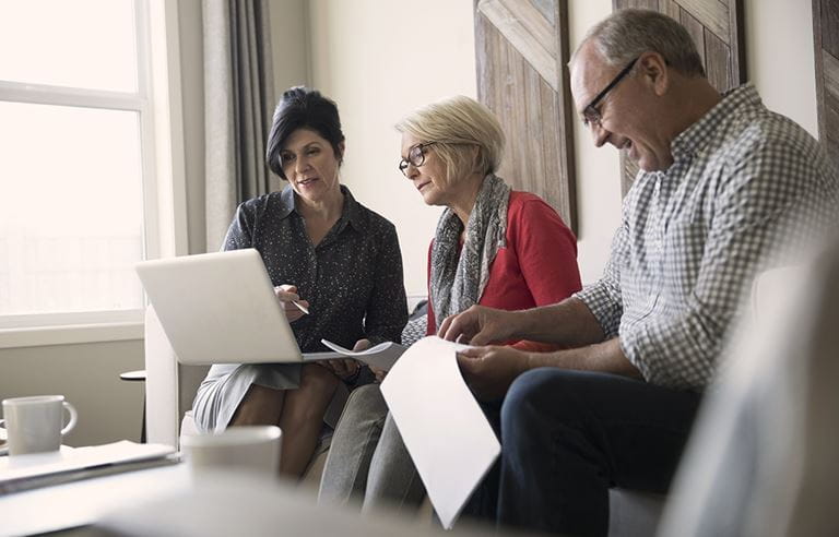 A woman is sitting with an elderly couple as they review her laptop and paperwork in their hands.