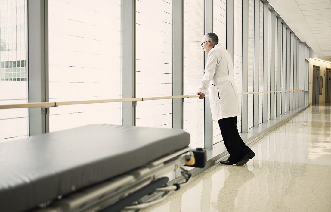 Doctor standing on a bridge connecting two hospital buildings looking out a window.