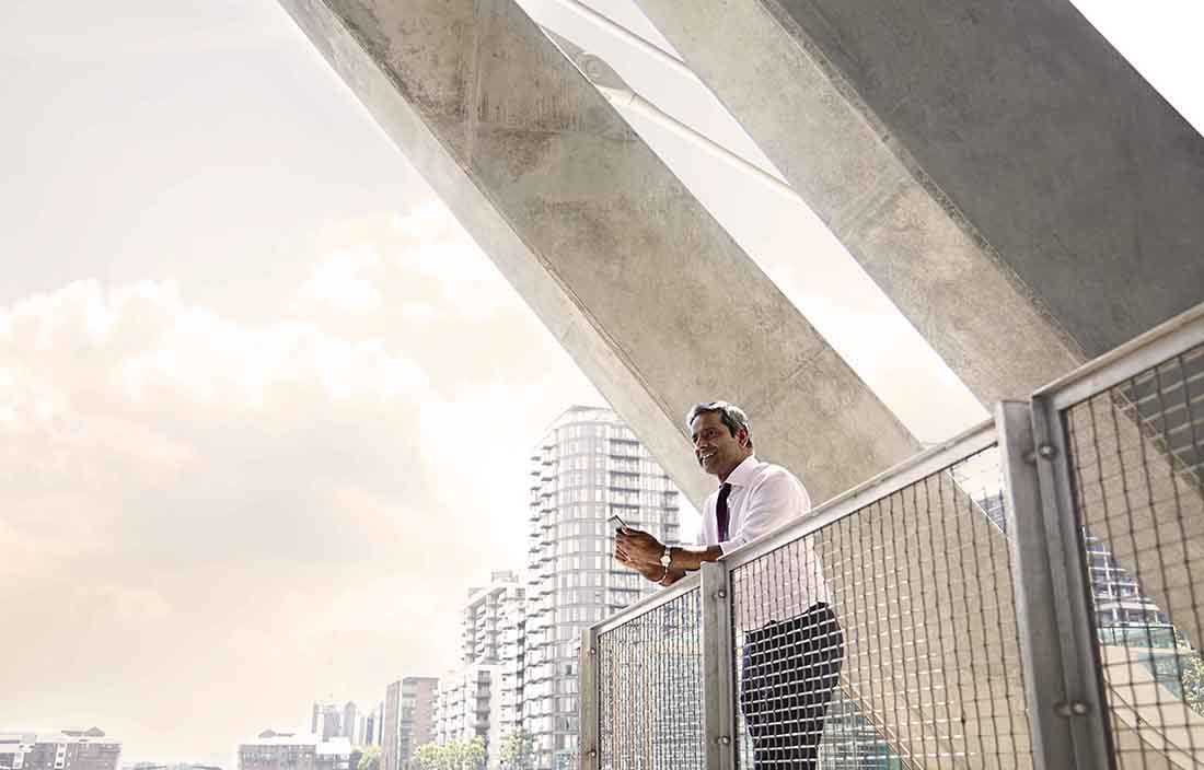 Businessperson standing in an outdoor area leaning on a railing while holding his cell phone.