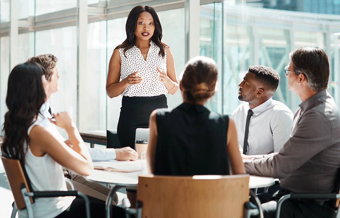 A group of business professionals having a meeting around a round table with one of them standing and speaking.